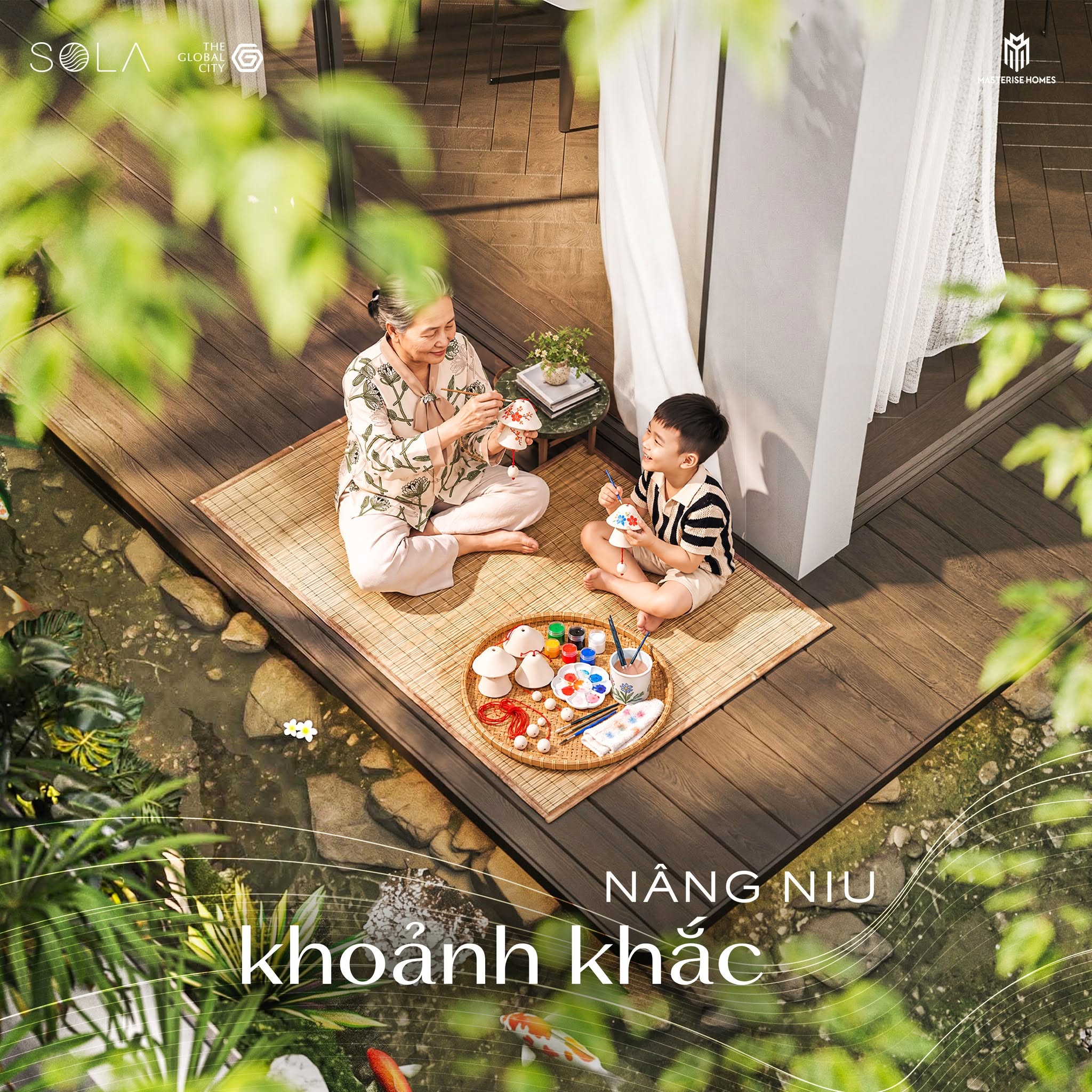 Một người phụ nữ lớn tuổi và một bé trai ngồi trên chiếu tre bên hồ cá koi, đang vẽ tranh. Cảnh được chụp từ trên cao, cho thấy không gian sân vườn và hiên nhà.