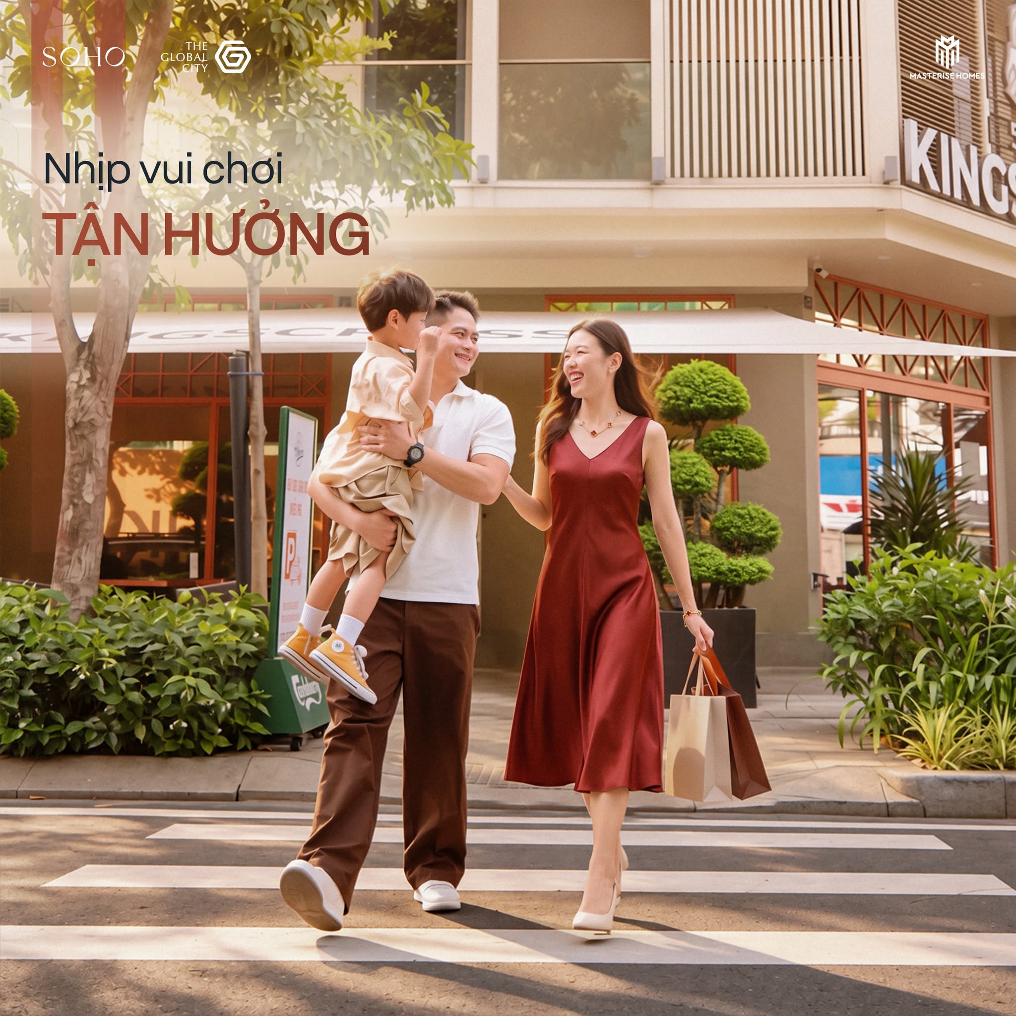 Ảnh chụp một gia đình gồm bố, mẹ và con nhỏ đang đi bộ qua vạch kẻ đường, người mẹ cầm túi mua sắm. Phía sau là các tòa nhà hiện đại với cửa hàng. Các logo SOHO, THE GLOBAL CITY và