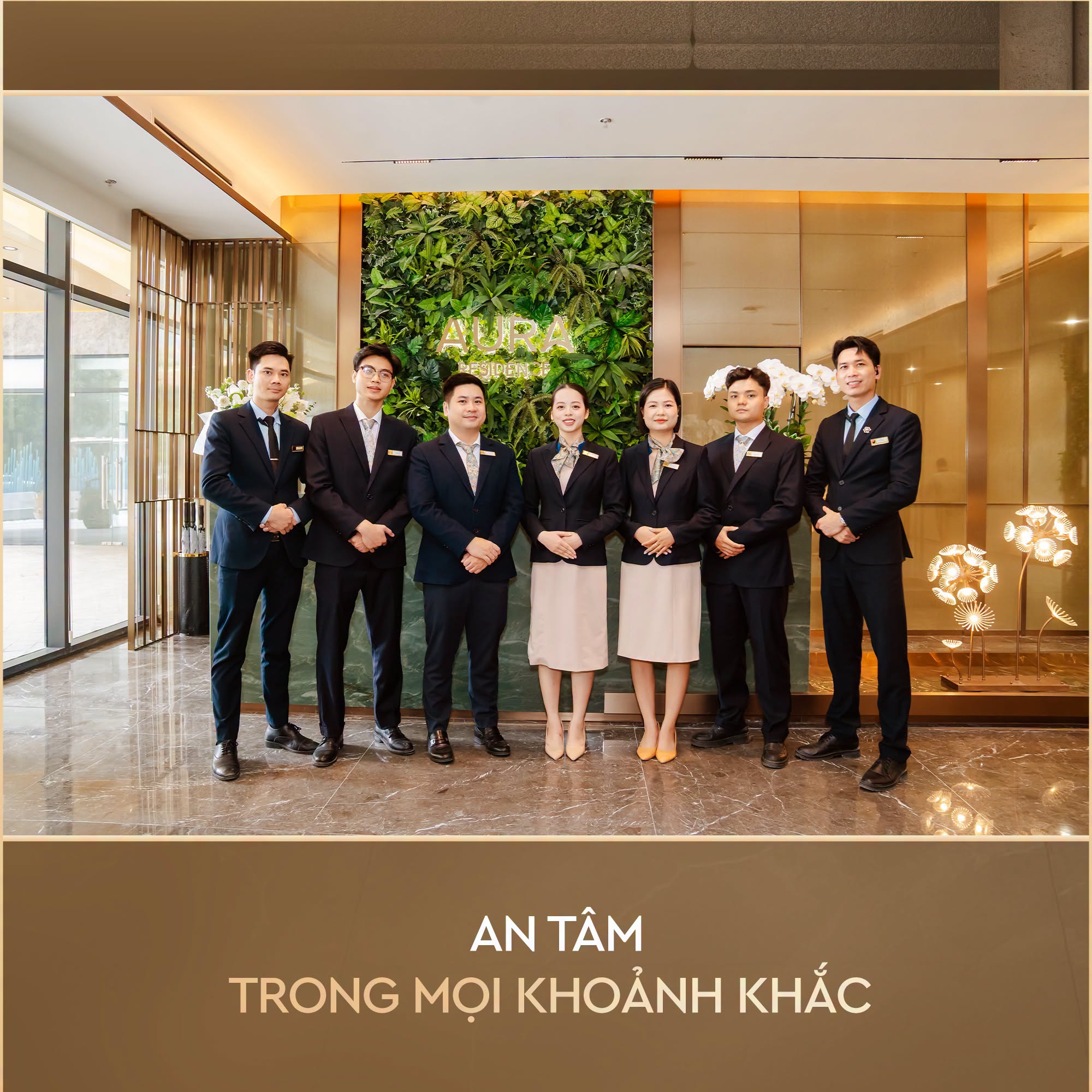 Nhóm 7 nhân viên (một số mặc vest, một số mặc đồng phục) đứng trong sảnh lễ tân hiện đại với bức tường cây xanh và chữ "AURA".